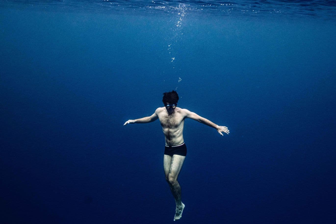 Ein Mann in Badehose und Schwimmbrille treibt ruhig im tiefblauen Meer, während über ihm eine Spur von Blasen aufsteigt.