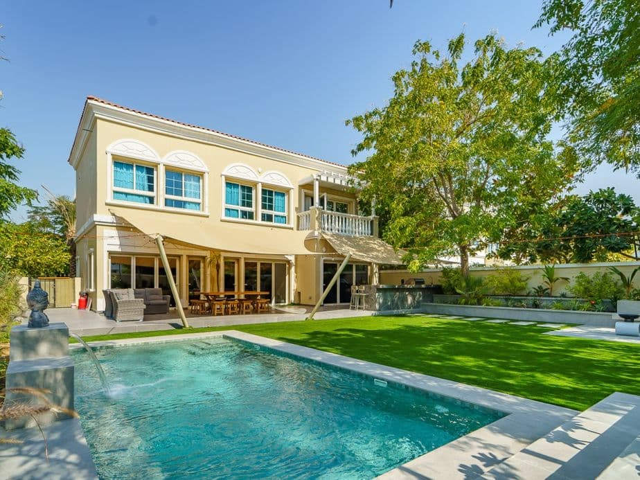 A two-story house with a large backyard featuring a swimming pool, patio with seating, and lush greenery under a clear blue sky.