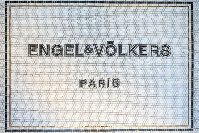 Ein Schild aus Mosaikfliesen mit der Aufschrift „ENGEL & VÖLKERS PARIS“ und einem schwarz-goldenen Rand auf weißem Hintergrund.