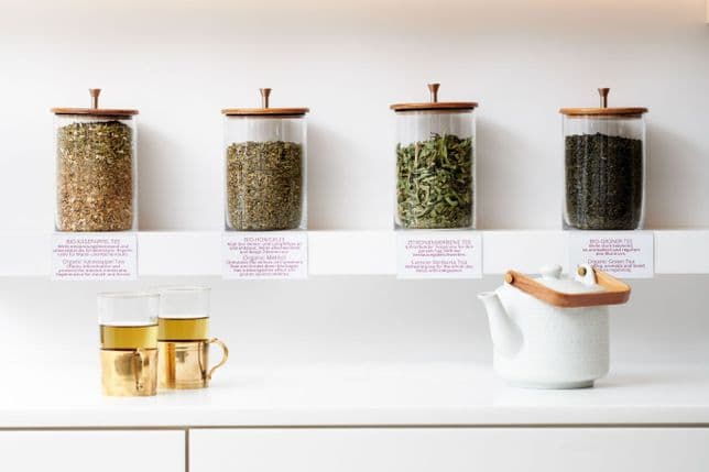 White tea shelf with four large glass containers with wooden lids, filled with loose herbs (cheese poplar, honey clover, lemon verbena, green tea). In front of it are two tea glasses with golden holders and a white teapot with a wooden handle.