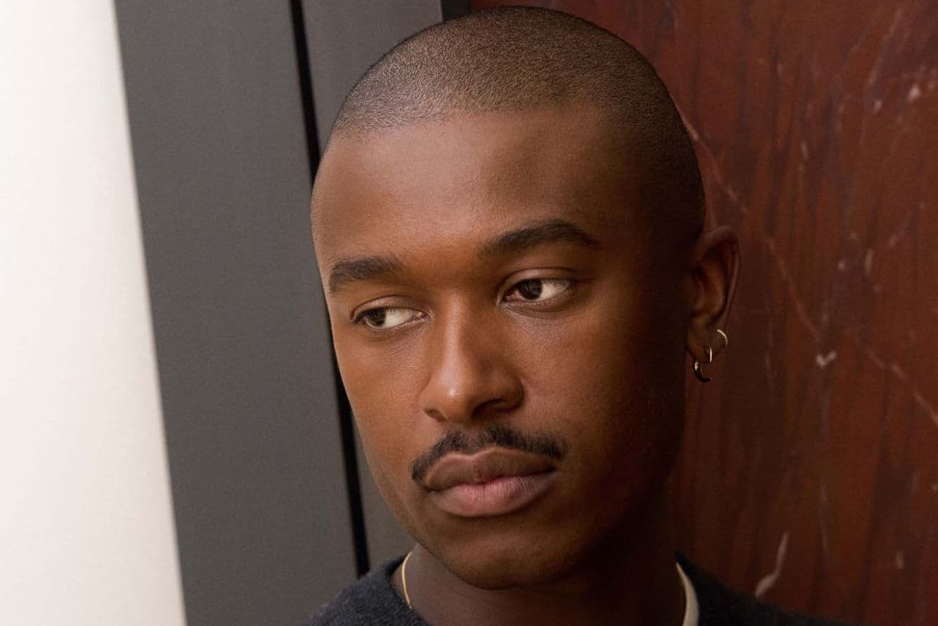 A person with a shaved head and earrings looks thoughtfully to the side, standing in front of a marbled wall.
