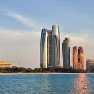 A skyline of modern skyscrapers, including reflective towers, stands along the waterfront under a clear blue sky.