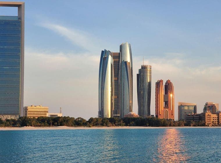 A skyline of modern skyscrapers, including reflective towers, stands along the waterfront under a clear blue sky.