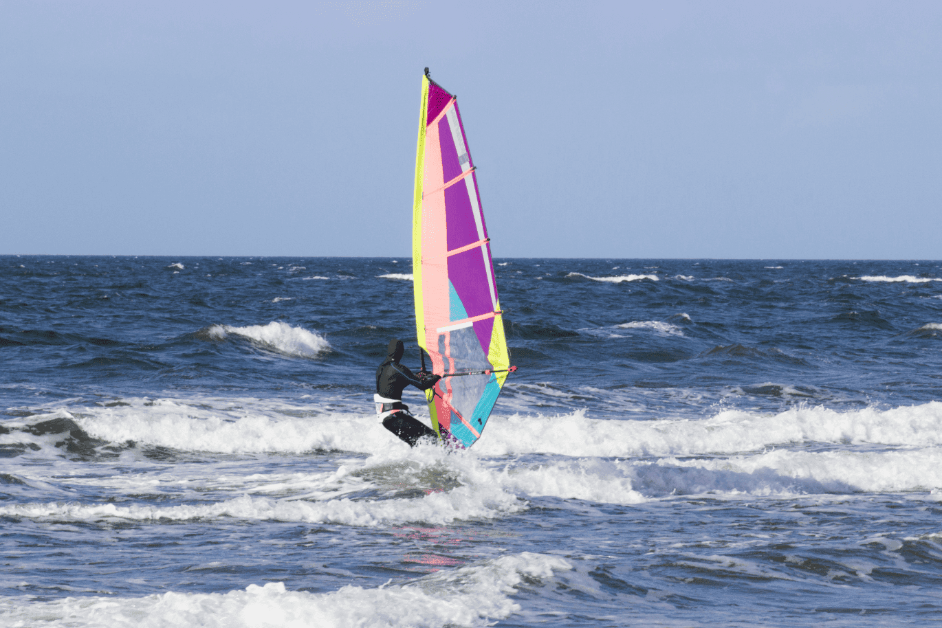 A person windsurfing on the ocean with a colorful sail, navigating through small waves under a clear blue sky.