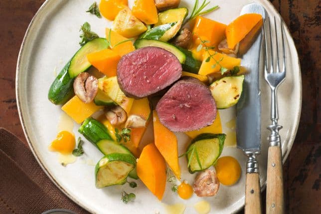 Light-coloured plate with two slices of pink beef fillet on roasted courgette and pumpkin vegetables, served with brown mushroom pieces, orange purée and fresh herbs; knife and fork on the right.