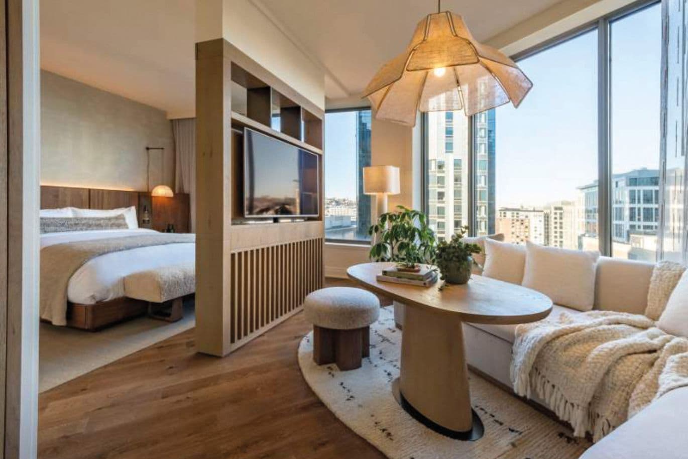 Bright hotel suite with partition TV wall, round table with plants, cream sofa, and bed in the background; city skyline visible through large windows.