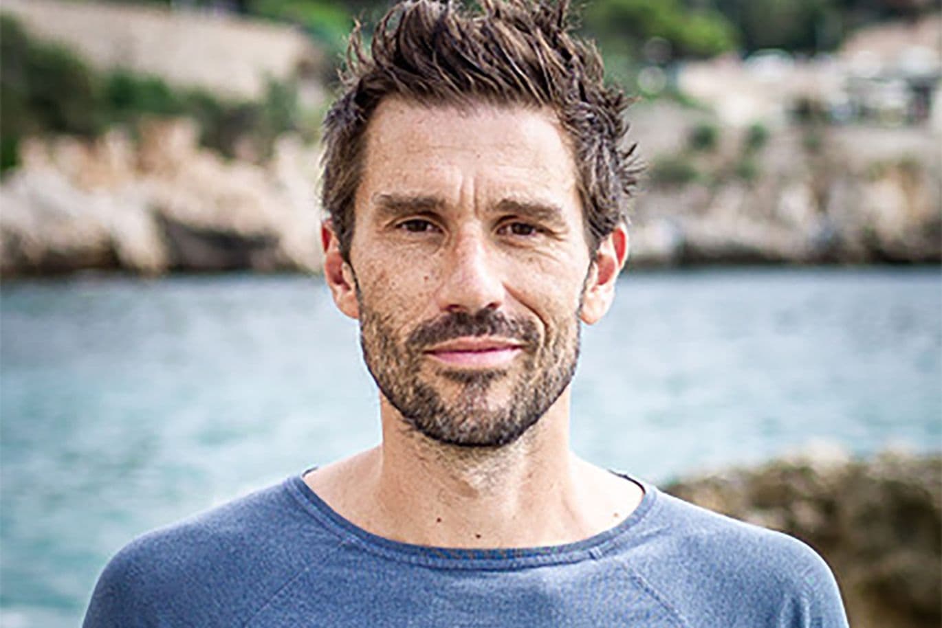 Ein Mann mit kurzen Haaren und Bart, der ein blaues Hemd trägt, steht im Freien vor einem verschwommenen Hintergrund aus Wasser und Grün.