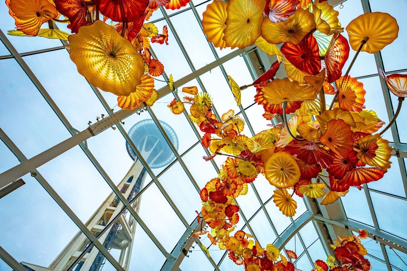 Vibrant orange and yellow glass flowers hang from a glass ceiling, with the Seattle Space Needle visible through the skylight.
