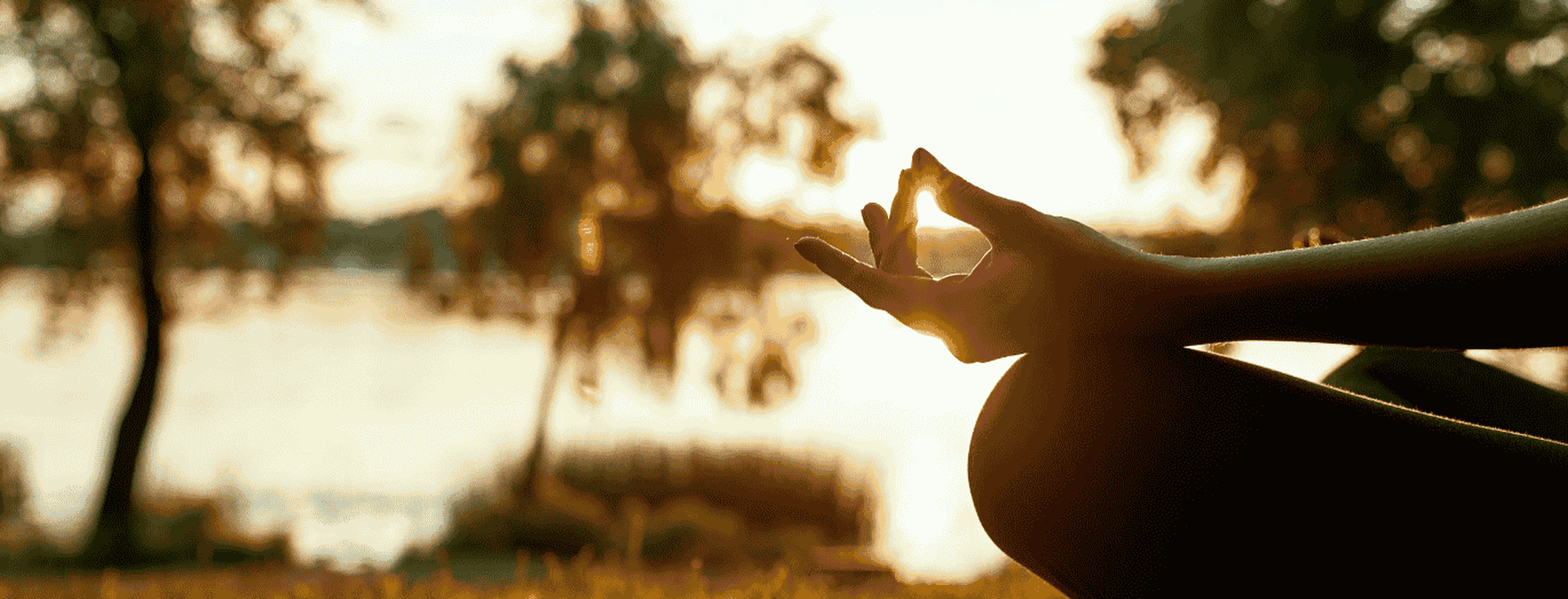 Person meditating outdoors at sunset by a lake, sitting cross-legged on a mat with hands in a mudra position.