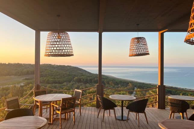 Eine Terrasse aus Holz am Meer mit runden Tischen, Korbstühlen und Hängelampen, von der aus man bei Sonnenuntergang den Blick auf grüne Hügel und das ruhige Meer genießen kann.
