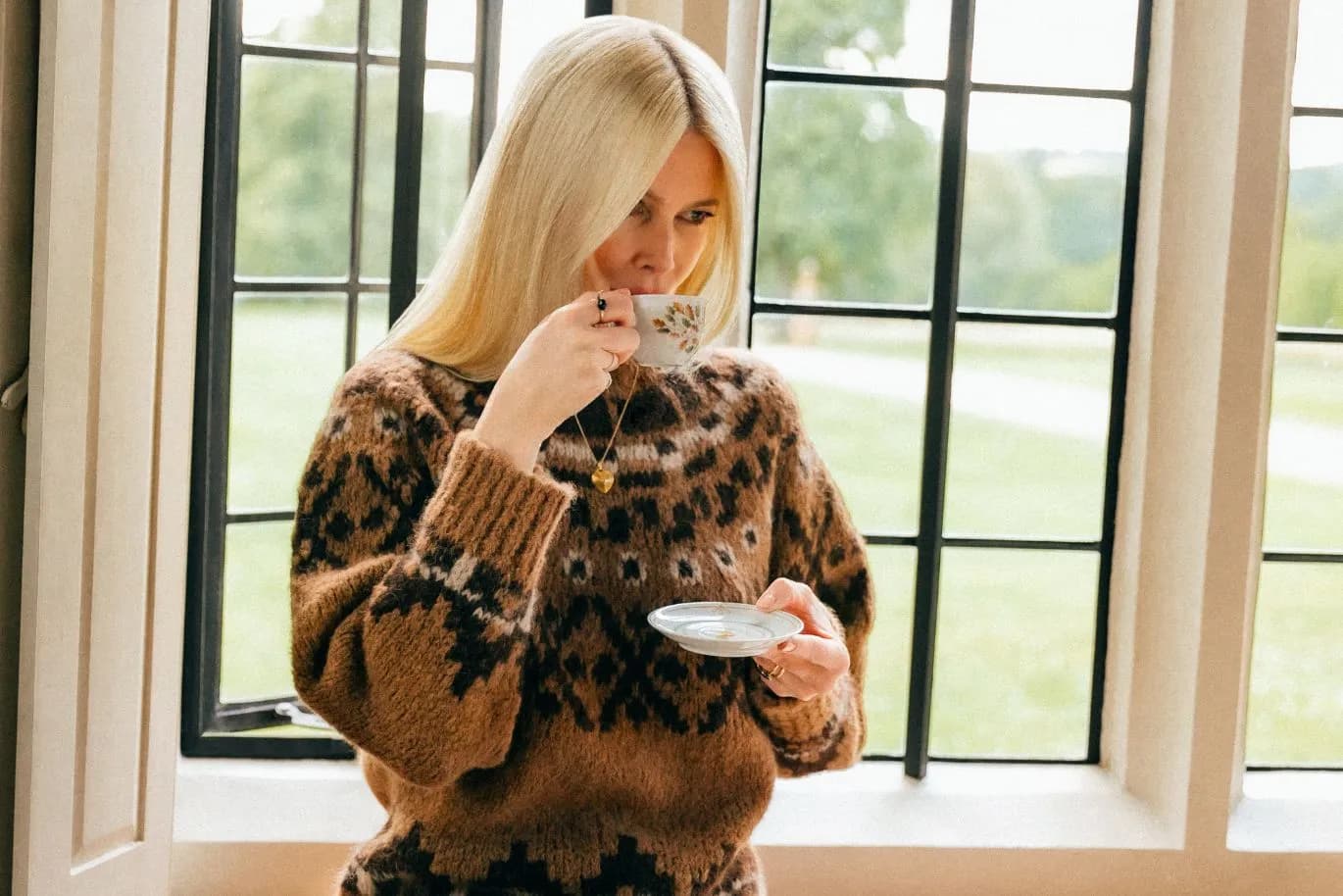 A blonde woman in a brown patterned sweater stands by a window, sipping from a teacup and holding a saucer.