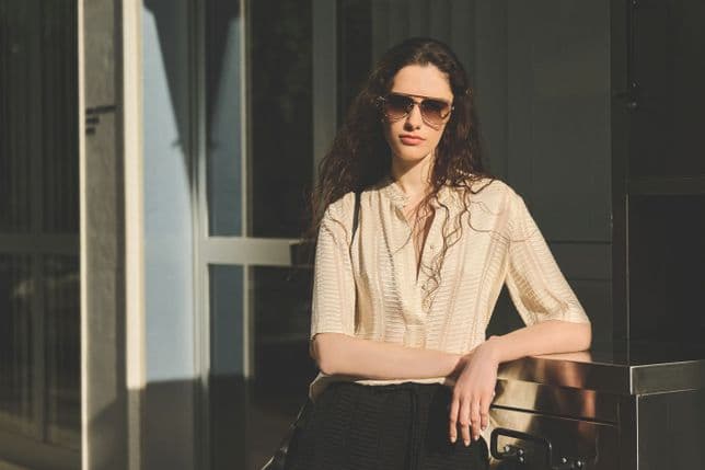 A woman wearing sunglasses, leaning against a metal surface, bathed in warm sunlight, with a modern building in the background.