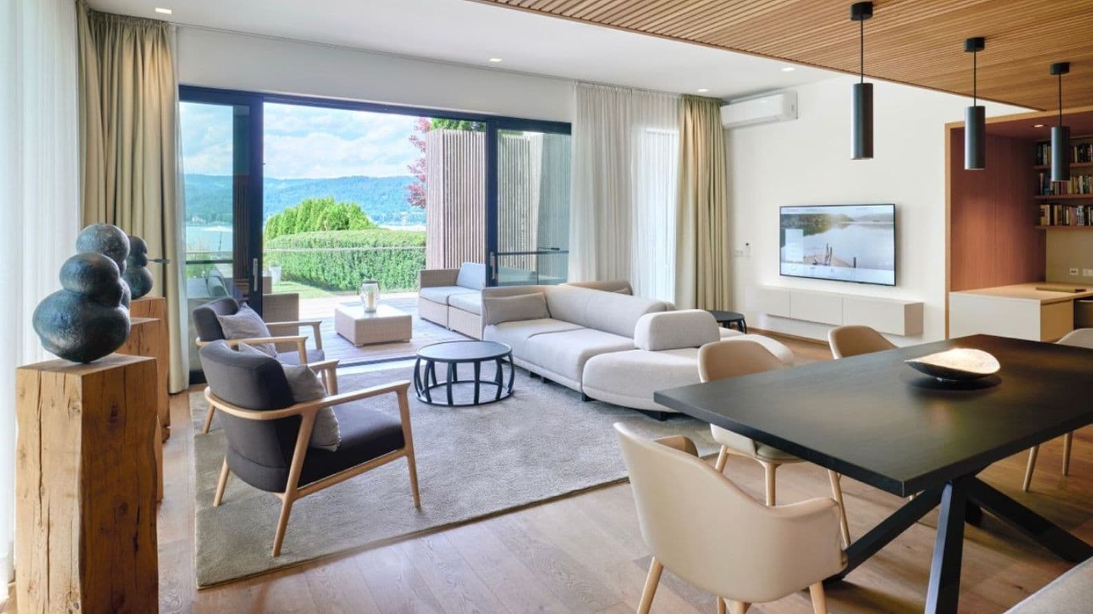 Modern, light-filled living and dining room; floor-to-ceiling sliding doors leading to the terrace with lake views, light-coloured sofas, black pendant lights, wooden slatted ceiling, wall-mounted TV, sculptures on wooden pedestals.