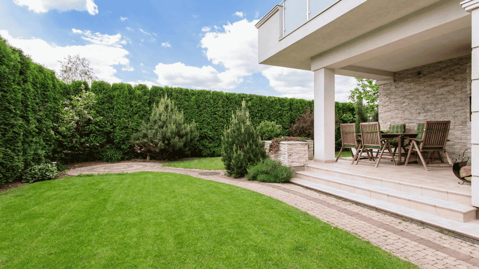 Ein weitläufiger Garten mit grünem Rasen, hohen Hecken und einer Terrasse mit einer Essgruppe unter dem Vordach eines modernen Hauses. Darüber erstreckt sich ein blauer Himmel mit weißen Wolken