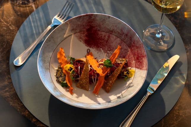 Elegant plate with crispy orange tuile, shredded vegetables, and herbs; burgundy-rimmed plate, wine glass, and cutlery on a blue placemat.