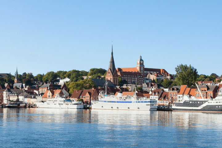 A scenic view of Flensburg's waterfront with boats docked, historic buildings, and a tall church tower under a clear blue sky.