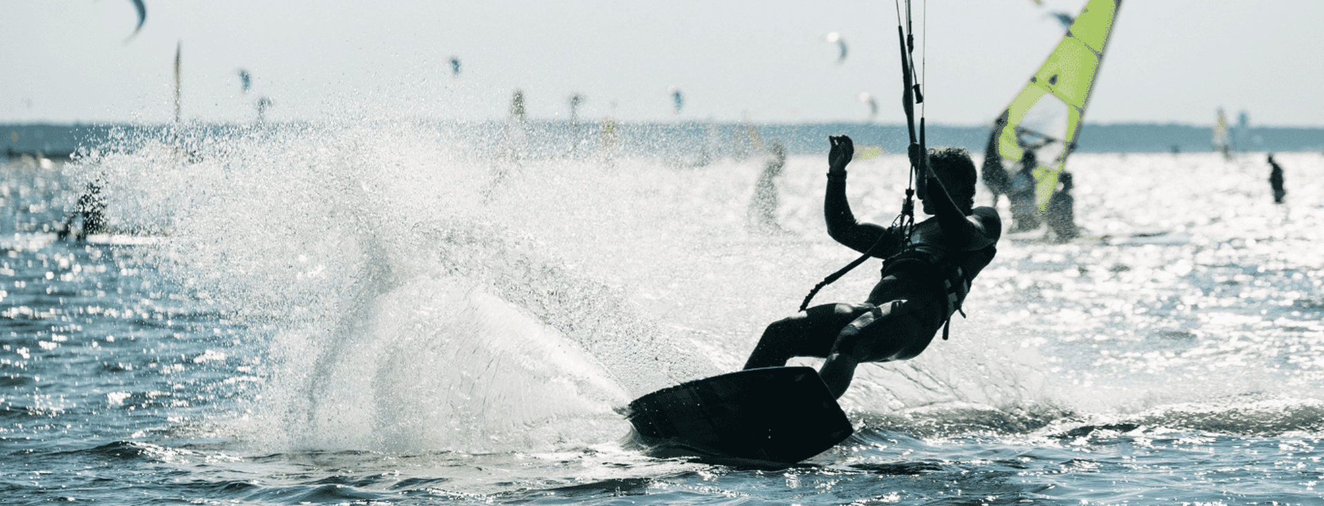 A person kiteboarding on a sunny day, creating a splash in the water, with multiple kites and a sailboat in the background.