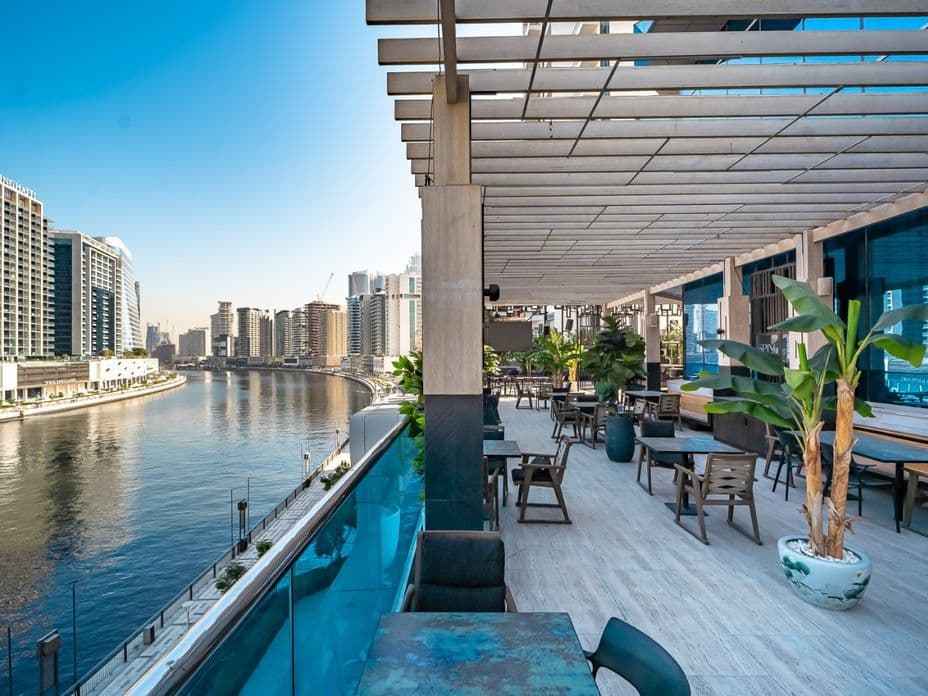 Riverside rooftop terrace with tables, potted palms and pergola, overlooking a curved canal and modern city skyline.