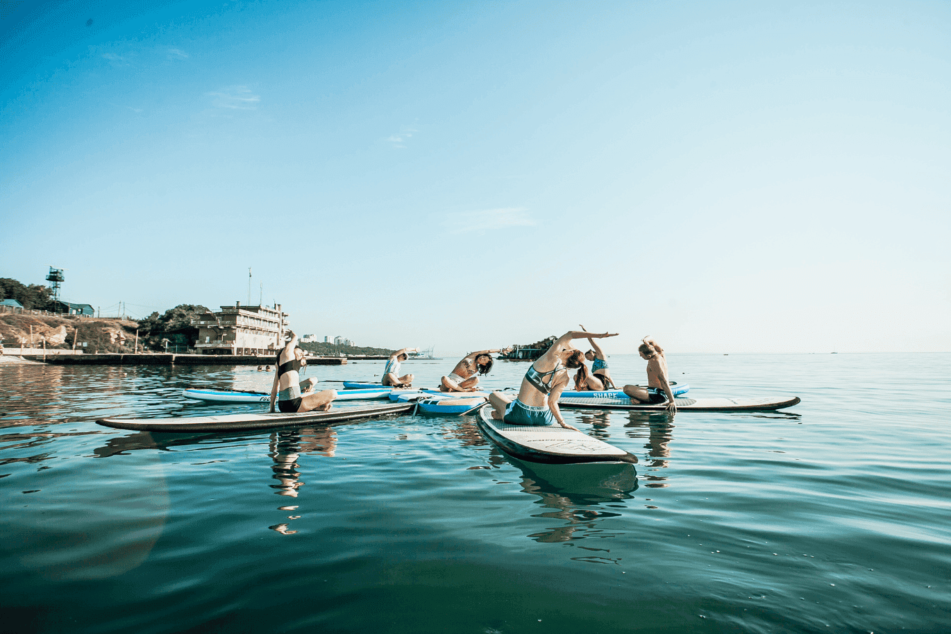 People practicing yoga on paddleboards in calm water near a dock, under a clear blue sky.