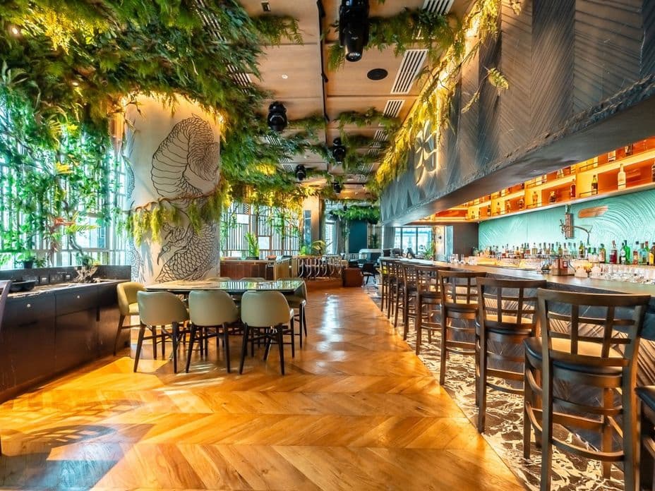 Bright upscale bar/restaurant interior with wooden herringbone floor, long bar and stools, lush hanging greenery, dragon mural column, warm lighting.