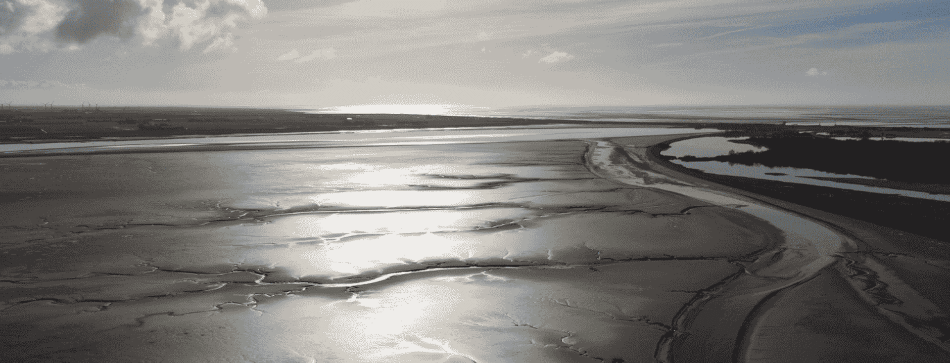 Sunlit coastal landscape with reflective wet sand, scattered clouds, and a winding shoreline under a bright sky.
