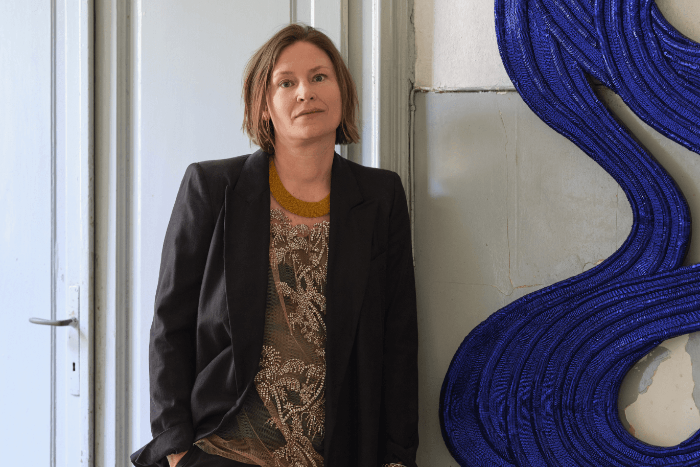 A woman with short brown hair, wearing a black blazer and patterned top, leans against a wall beside a blue coiled sculpture.