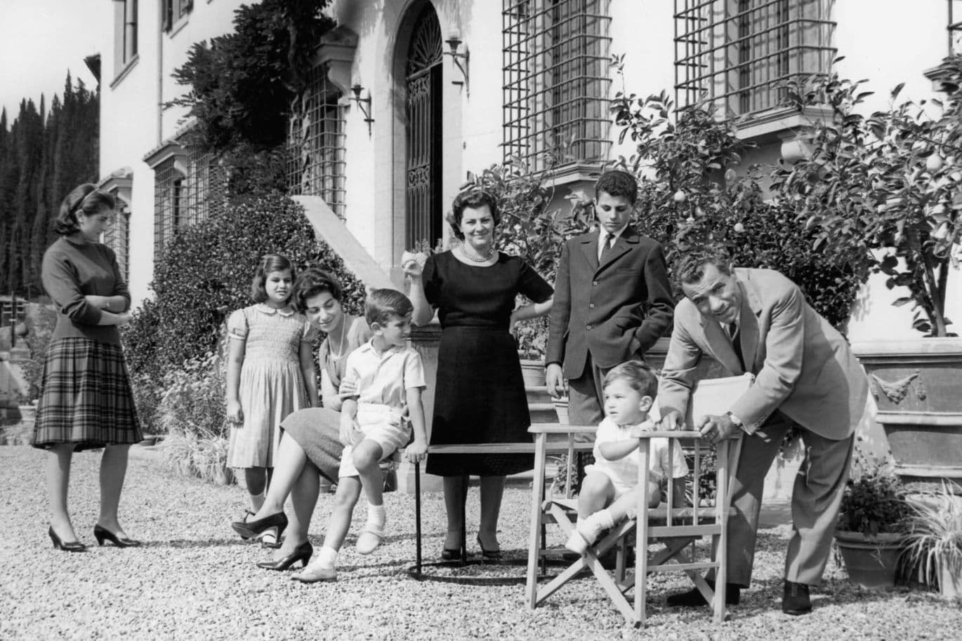 A family poses in front of a large house. Adults and children gather around a toddler in a high chair. The scene is formal and sunny.