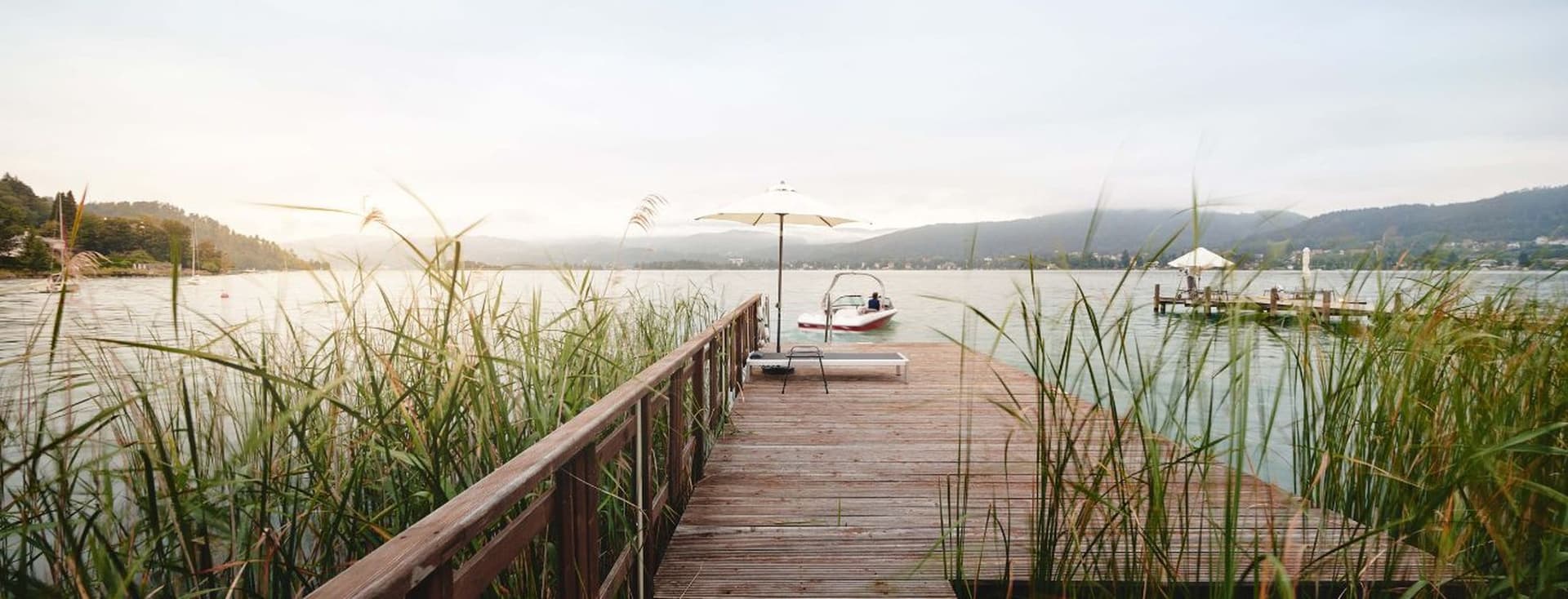 Wooden jetty through reeds on a tranquil lake; at the end, a sun lounger and a white parasol, next to it a moored motorboat. In the background, hills, the shoreline and another jetty in the soft evening light.