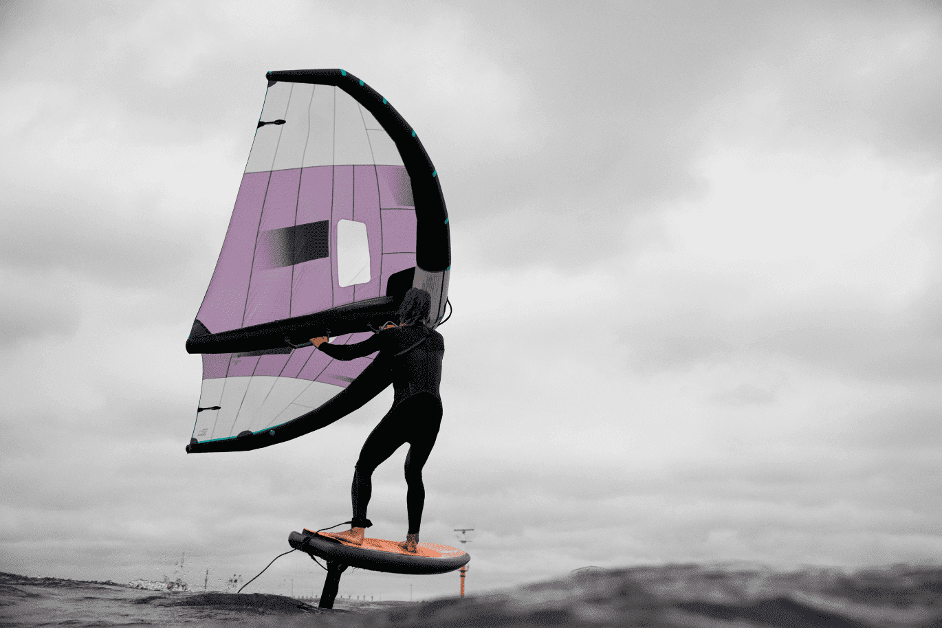 Person wing foiling on water under cloudy skies, holding a purple and white sail.