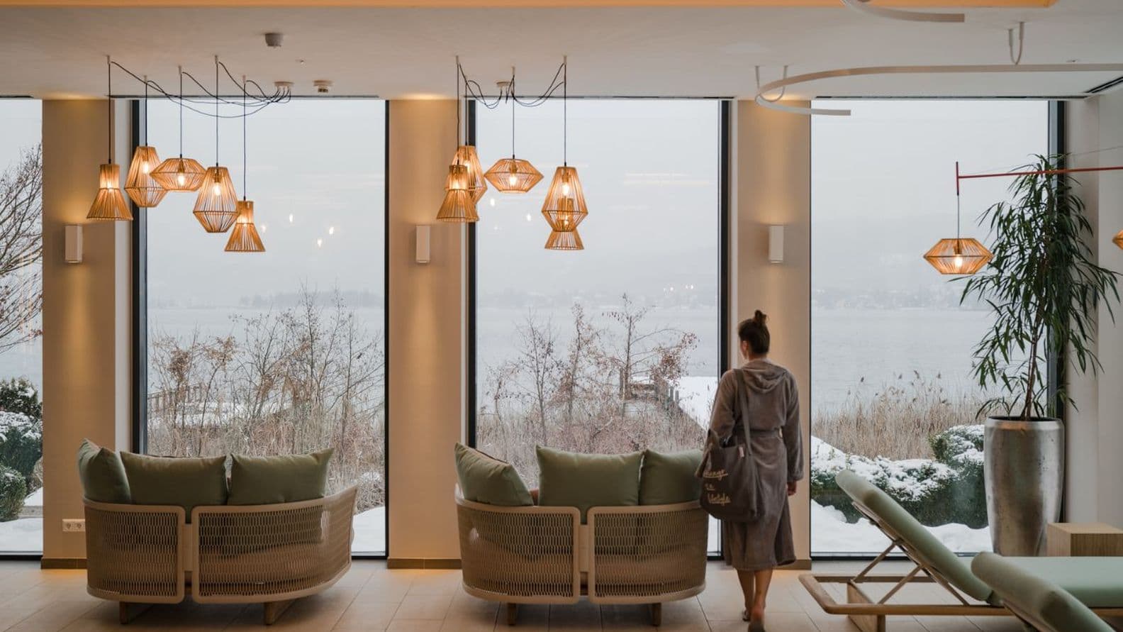 Bright spa lounge with large panoramic windows overlooking the snow-covered lake; sofas and loungers, warm pendant lights; person in grey bathrobe walks to the window.