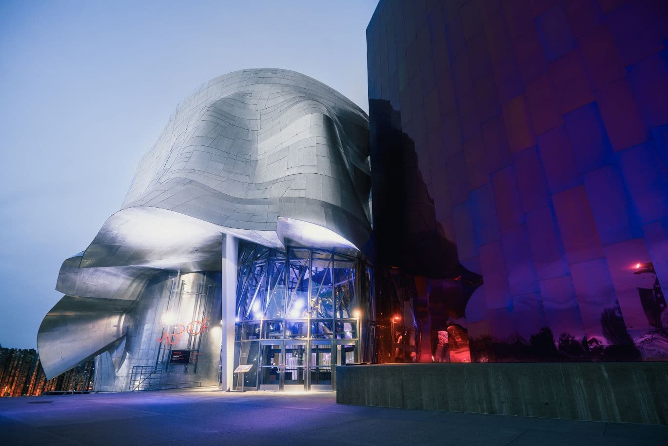 Nighttime shot of a futuristic silver building with a wavy metal facade and glass entry, beside a tall, purple-reflective wall.