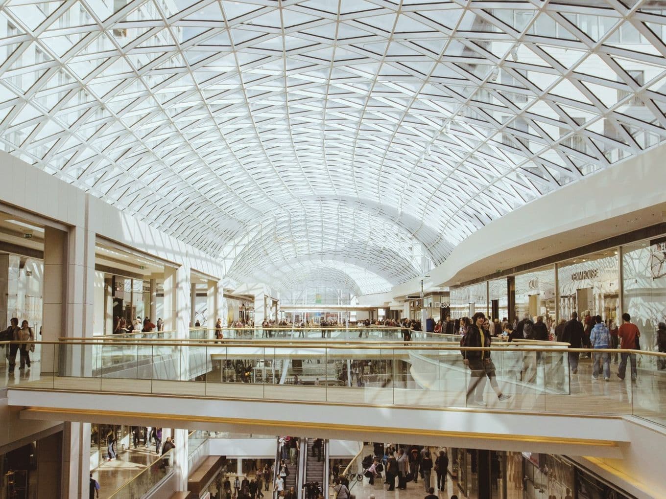 Spacious multi-level shopping mall with a white geometric glass arched ceiling, glass railings and crowds of shoppers.