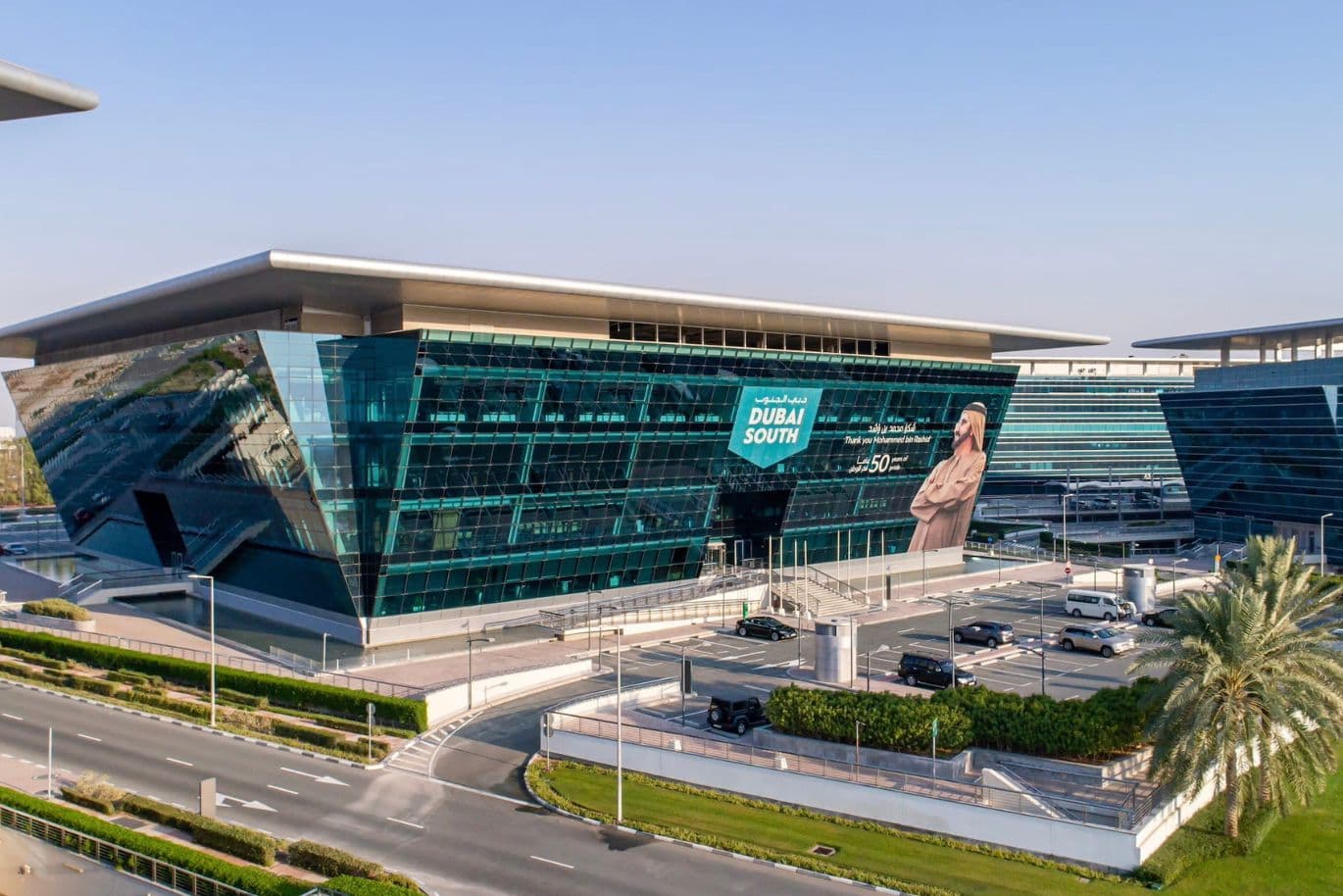 Modern glass building with angular design and a Dubai South banner; surrounding roads, cars, and a palm tree in a landscaped area.