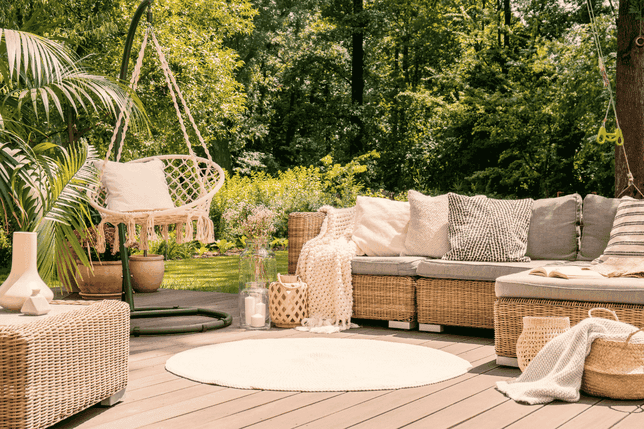 Eine gemütliche Terrasse mit Korbmöbeln, Kissen, einem Hängesessel und Pflanzen, die auf einem Holzsteg inmitten von Grün liegt