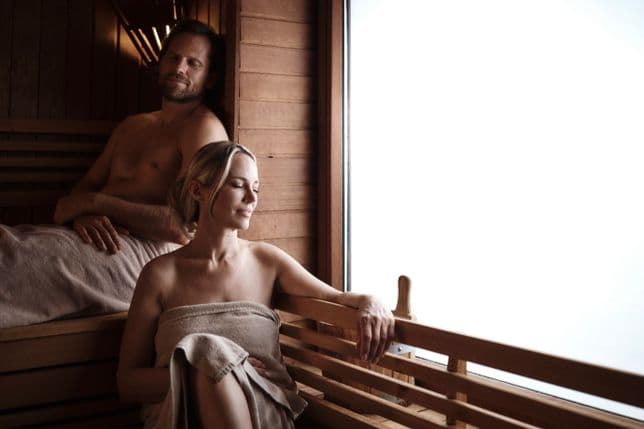 Two people are relaxing in a wood-panelled sauna; one person is sitting in front on the lower bench near a large, bright window, wrapped in a towel, while another is resting on the upper bench behind them.