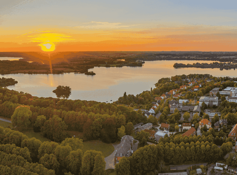 Luftaufnahme eines Sonnenuntergangs über einem ruhigen See, umgeben von üppigem Grün und einem kleinen Dorf, mit leuchtenden Orange- und Blautönen am Himmel