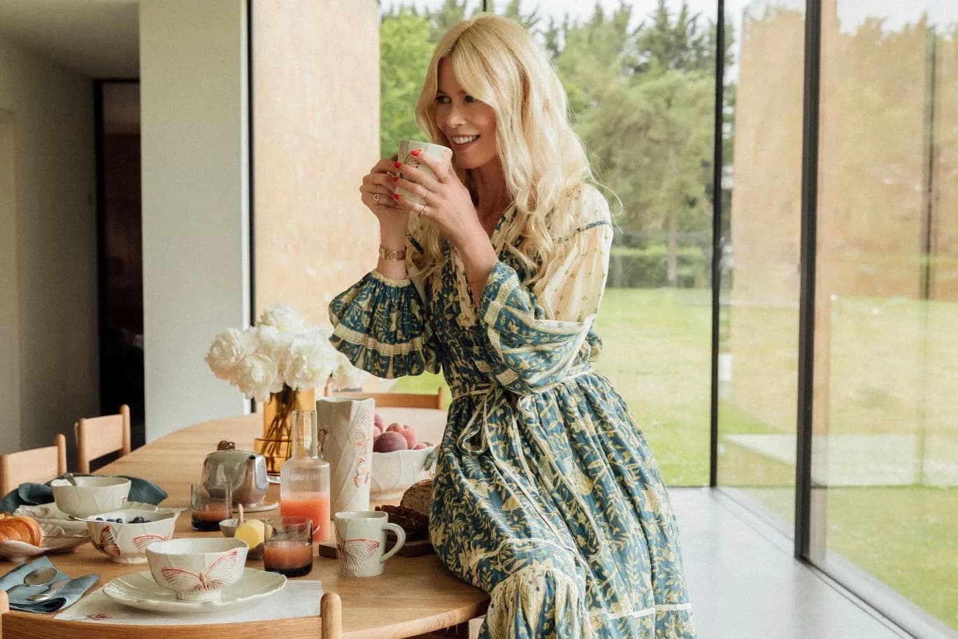 Smiling blonde woman in a floral dress sits at a breakfast table by large windows, holding a cup amid dishes and flowers.