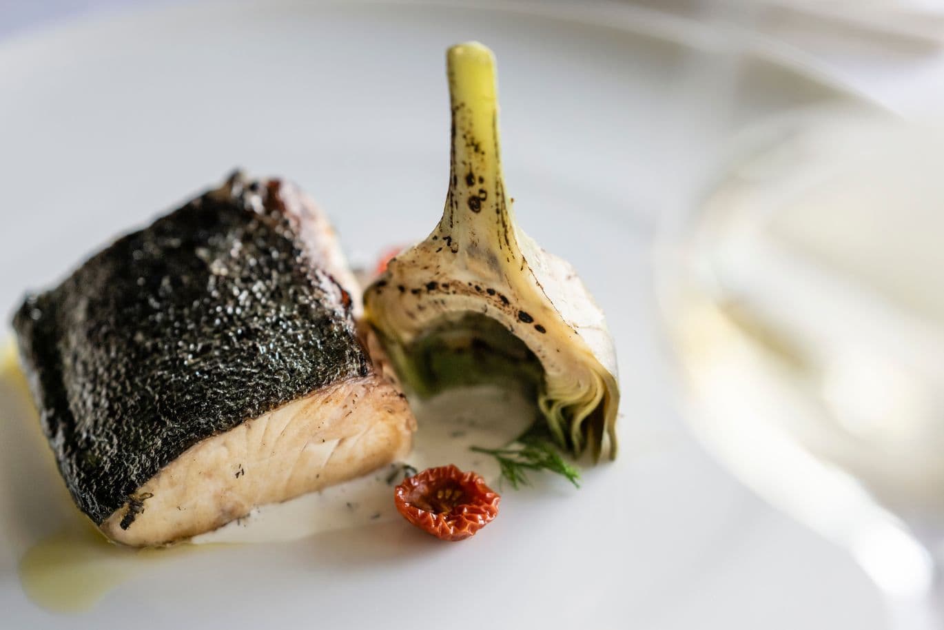 Gebratenes Fischfilet mit knuspriger Haut, einer angebratenen Artischockenhälfte und sonnengetrockneten Tomaten, serviert auf einem weißen Teller.