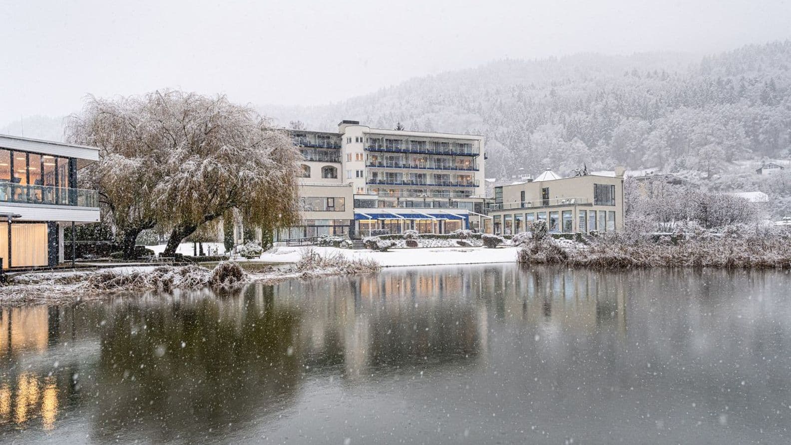 Hotel by the lake in heavy snowfall; snow-covered trees and hills in the background, warm window lights; calm water with reflections in the foreground.