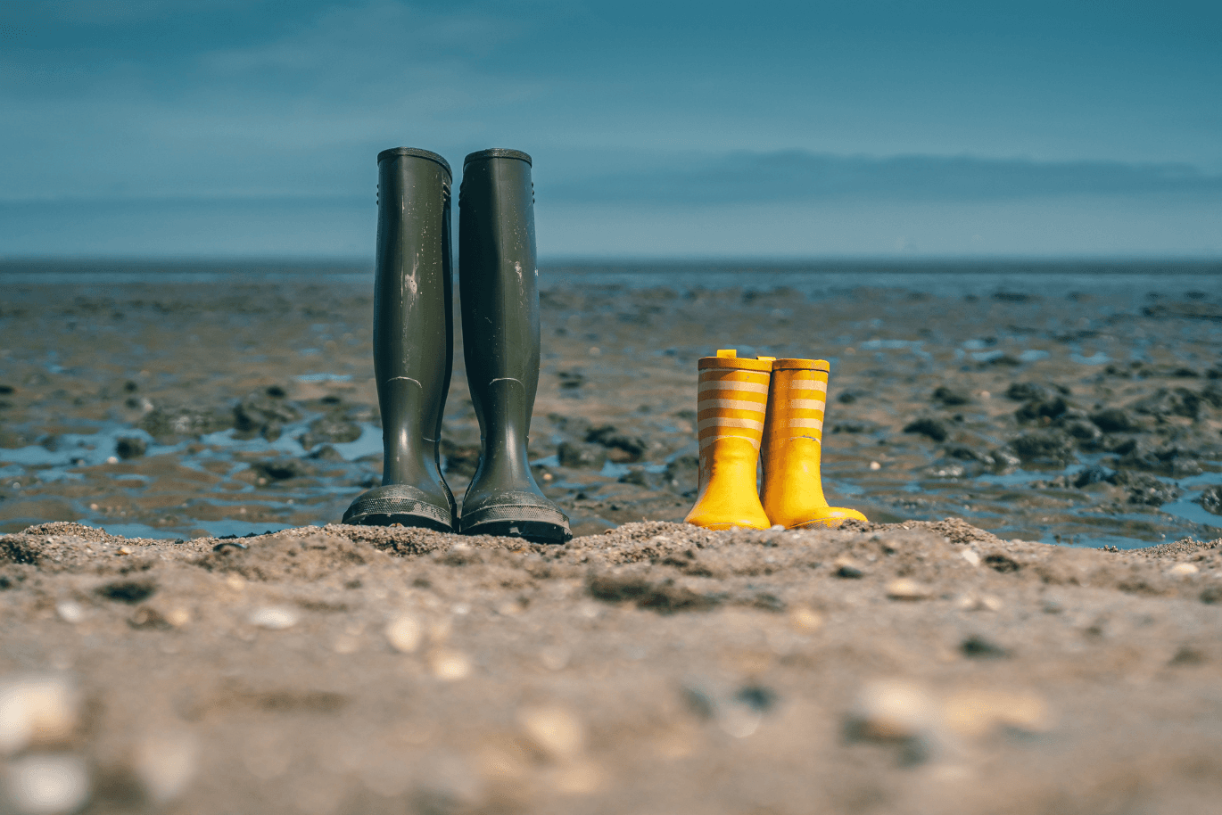 Two pairs of rubber boots, one green and one yellow with stripes, stand on a sandy beach with a rocky shoreline in the background.