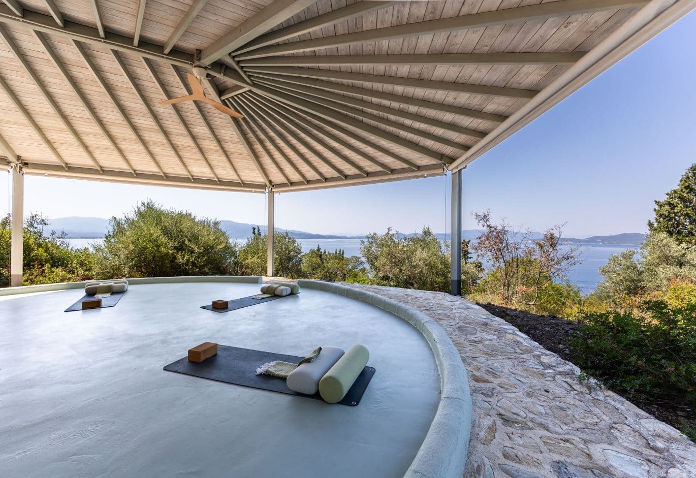 Open-air circular yoga pavilion with a wooden roof structure and laid-out mats, overlooking the sea.