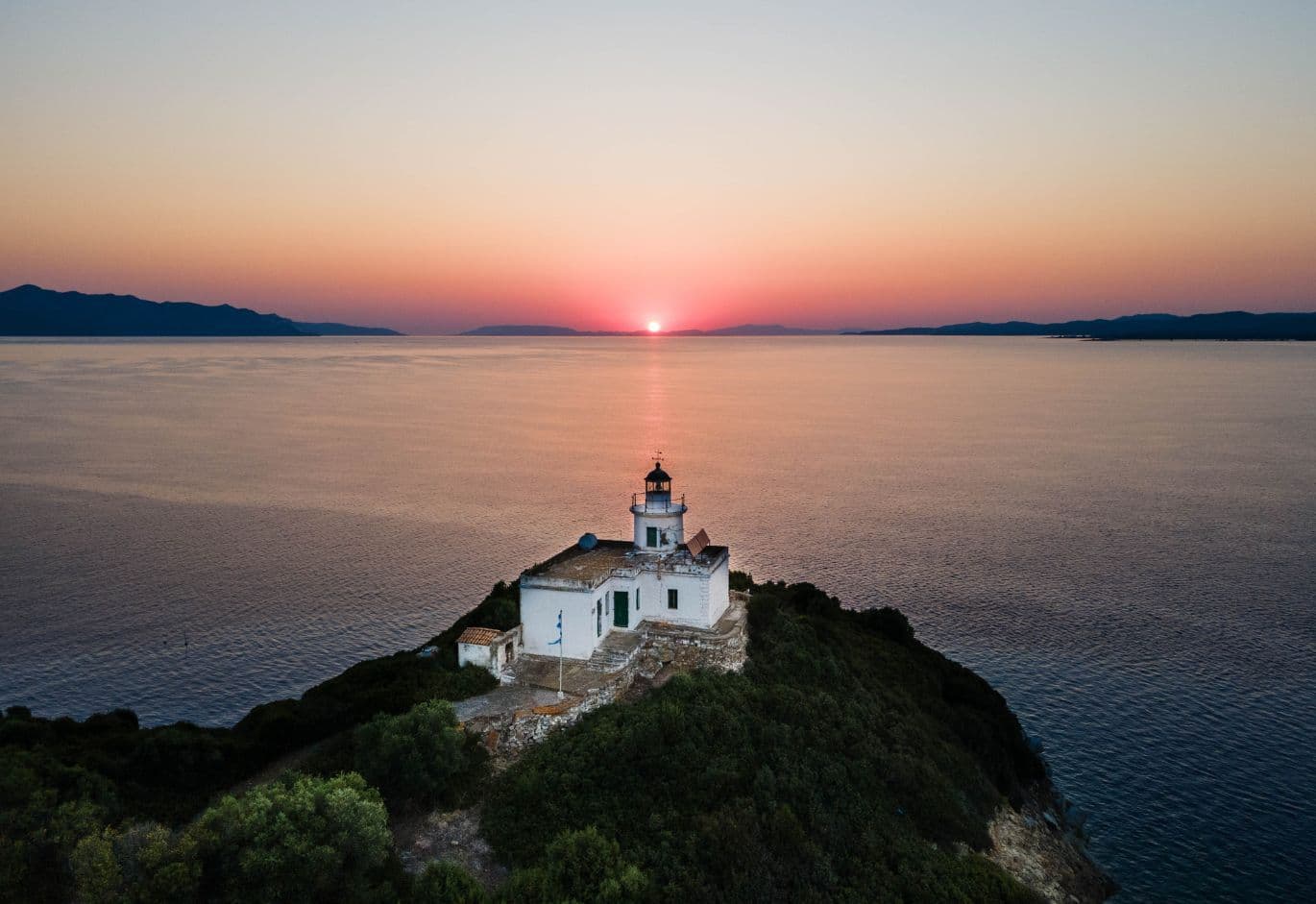 Aerial sunset view of a white lighthouse on the tip of a green peninsula overlooking calm waters.