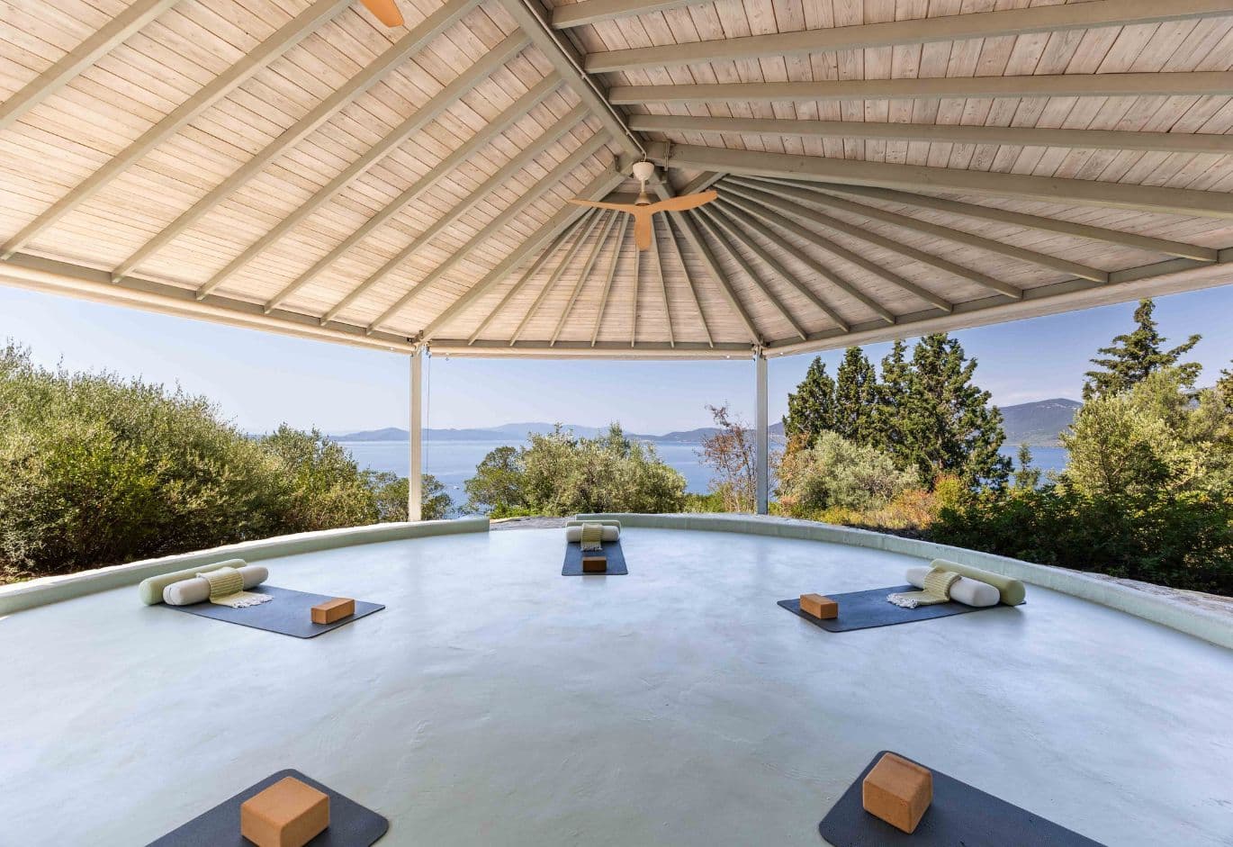 Open-air circular yoga pavilion with a wooden roof structure and laid-out mats, overlooking the sea.
