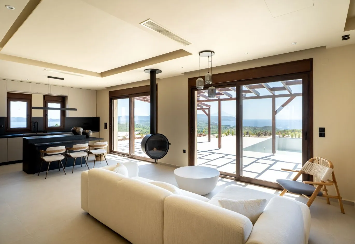 Modern open-plan interior with off-white sofa, suspended black fireplace, kitchen island, and large sliding glass doors leading to a terrace with sea views.