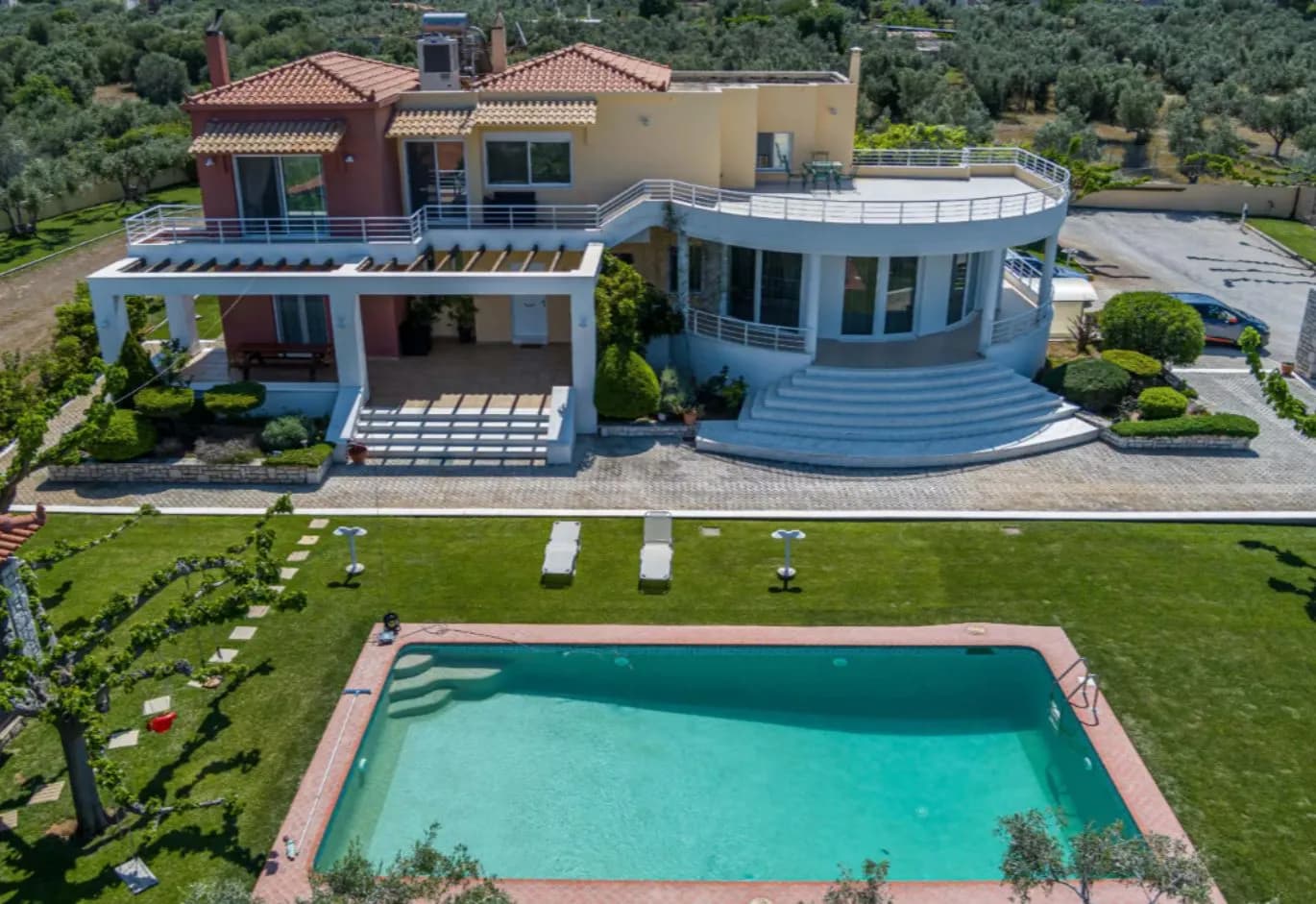 Aerial view of a luxury estate featuring a multi-level villa with a semi-circular balcony, large swimming pool, and sprawling green lawn surrounded by olive trees.
