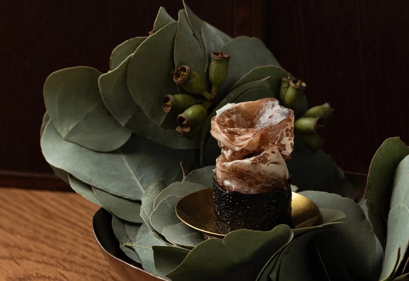 Artistic bite-sized gourmet dish wrapped in nori and thinly sliced meat, served over eucalyptus leaves