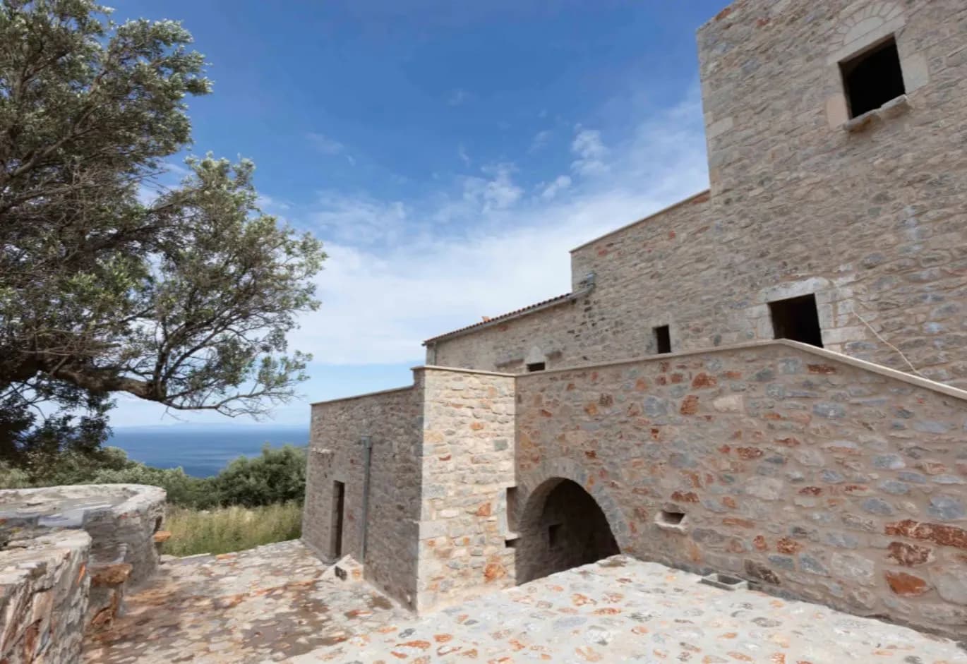 Traditional stone-built house with paved terraces, arched details, and an olive tree against the sea background.