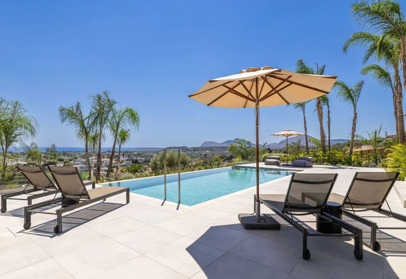 Luxurious outdoor pool area with sun loungers, umbrellas, palm trees, and a panoramic sea view.