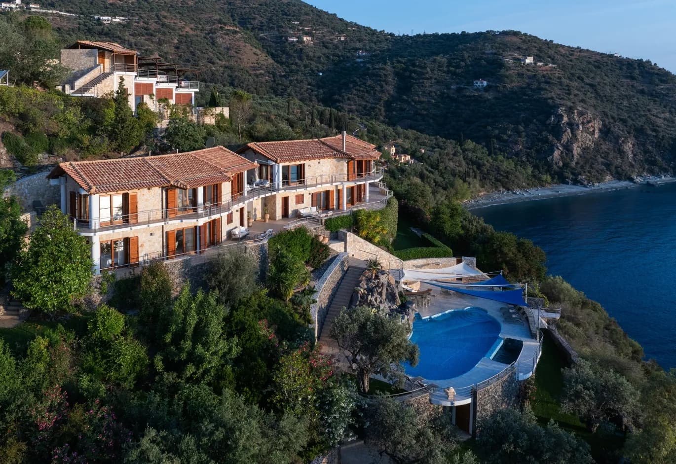 Panoramic aerial view of coastal villas nestled along a lush green hillside overlooking a tranquil sea cove.
