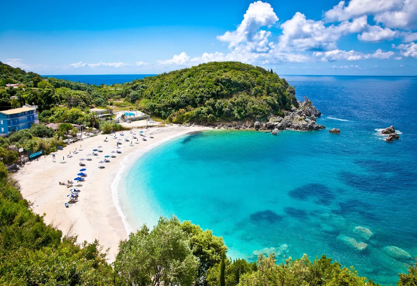 Aerial view of a turquoise bay and sandy beach framed by lush green hills, blue sea, and summer sky.